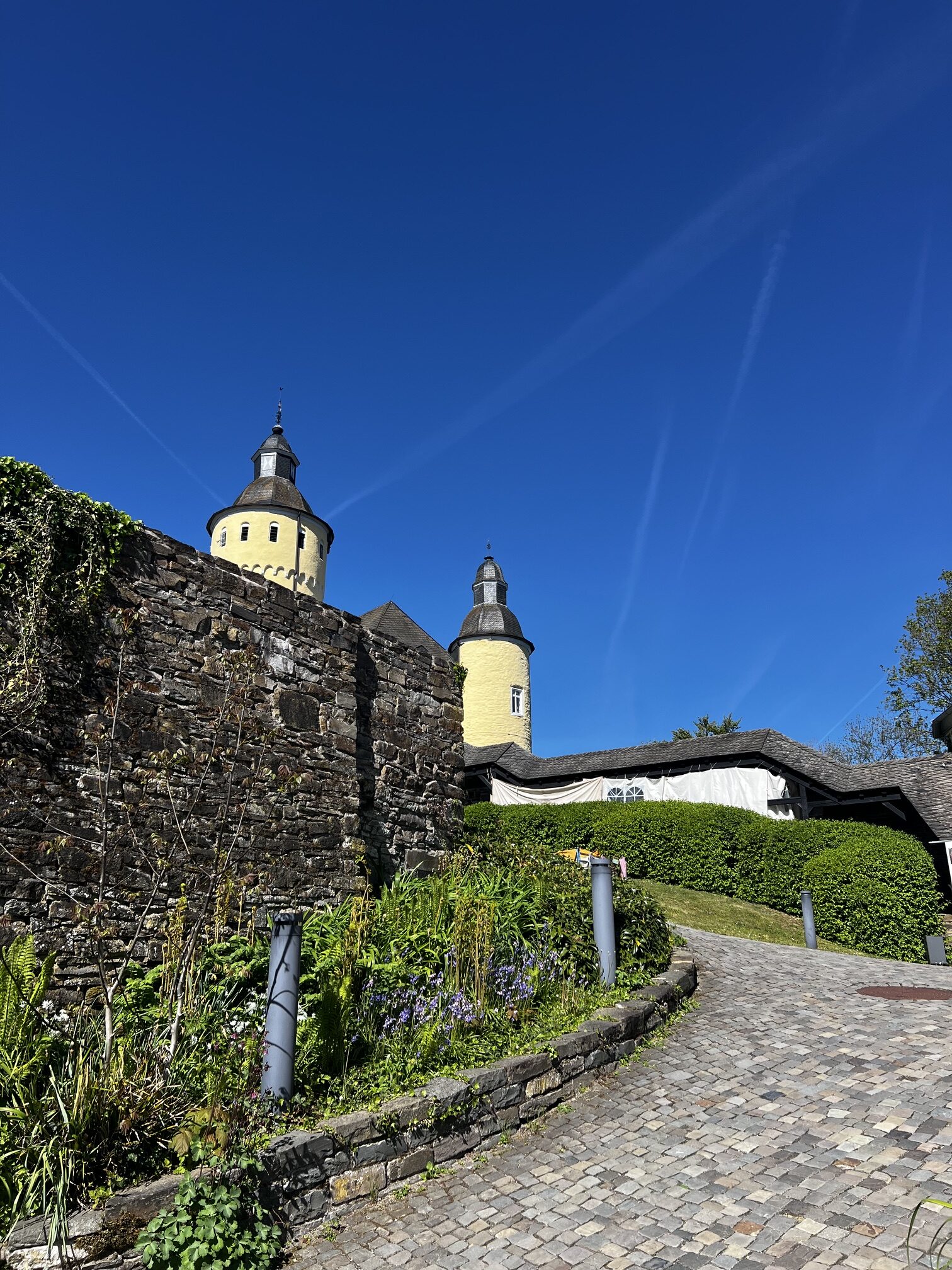 Ein kopfsteingepflasterter Weg führt an einer Steinmauer und Grünanlagen vorbei zu einem gelben Schloss mit zwei runden Türmen unter einem klaren blauen Himmel.