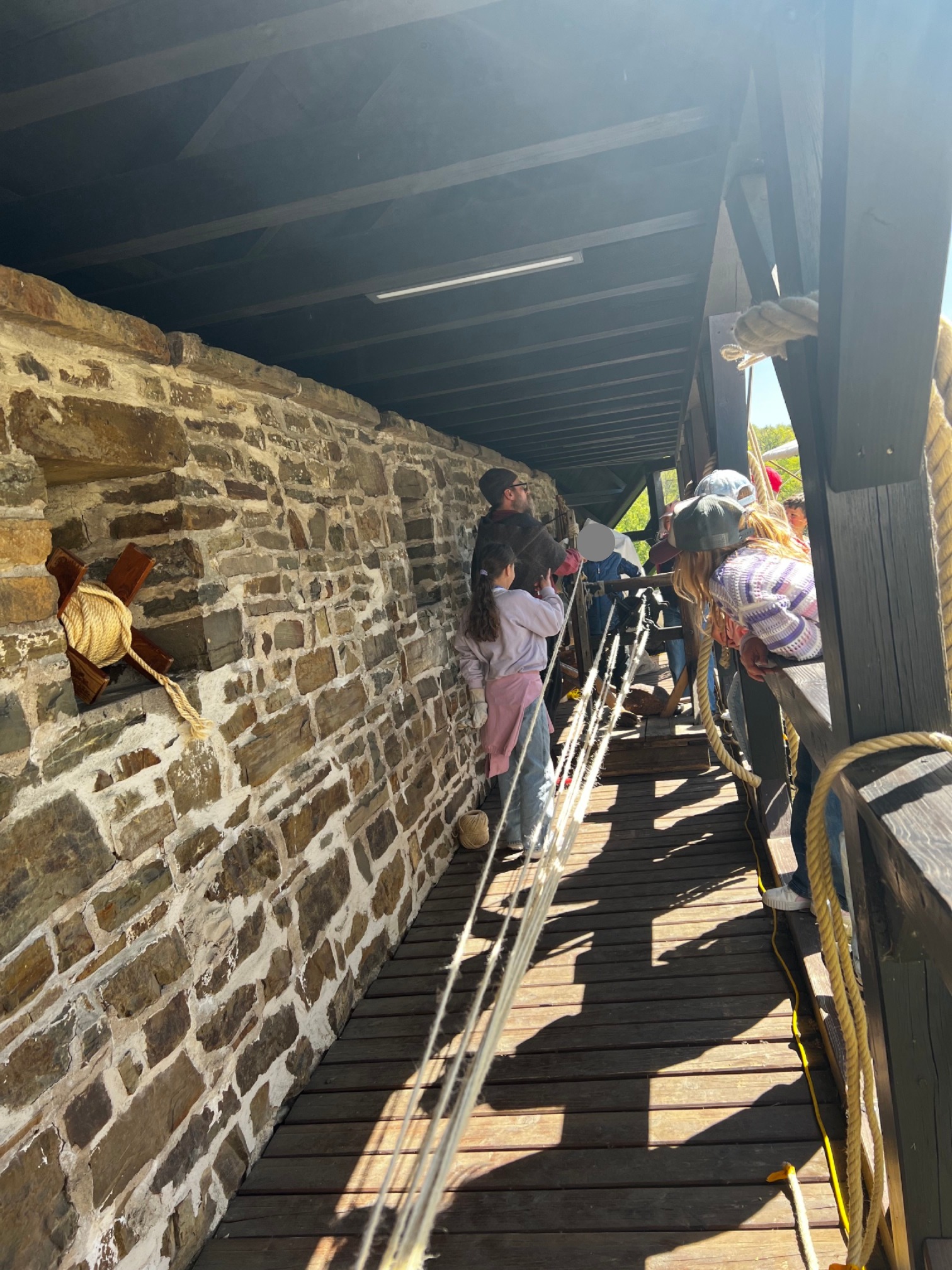 Menschen stehen und beobachten auf einer Holzbrücke neben einer Steinmauer, an der Seile befestigt sind und durch die das Sonnenlicht fällt.